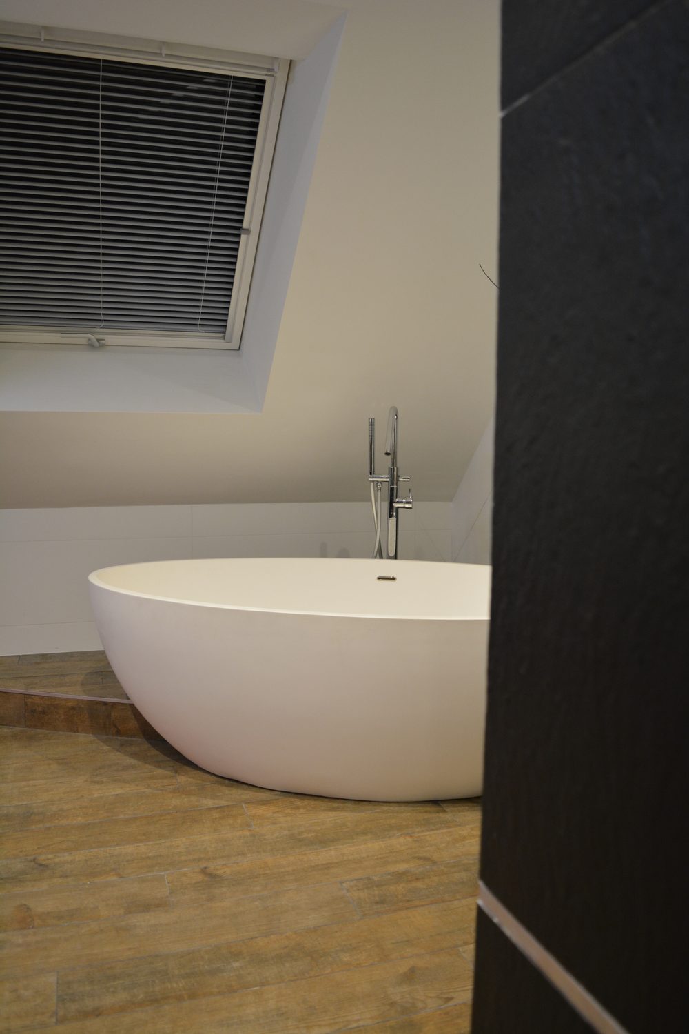Baignoire îlot ovale avec robinetterie sur pied sous une fenêtre de toit, sol carrelage imitation bois