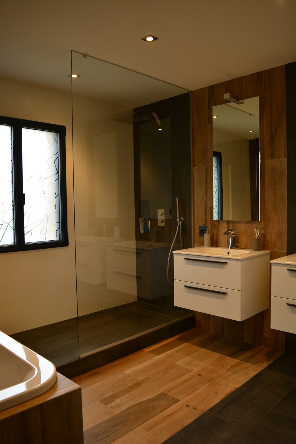 Vue d'ensemble de la salle de bain de Rezé : douche à l'italienne sur estrade avec paroi de verre, vasque suspendue, sol imitation parquet